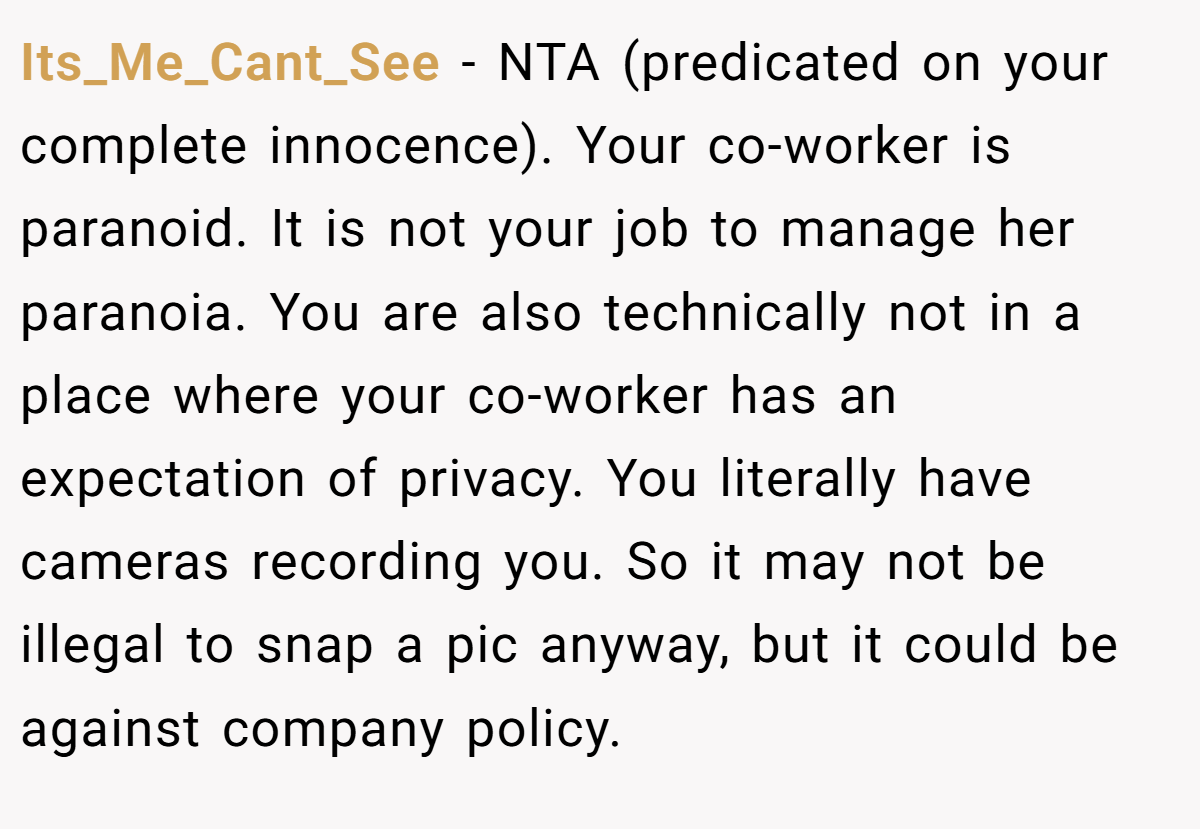 Its_Me_Cant_See − NTA (predicated on your complete innocence). Your co-worker is paranoid. It is not your job to manage her paranoia. You are also technically not in a place where your co-worker has an expectation of privacy. You literally have cameras recording you. So it may not be illegal to snap a pic anyway, but it could be against company policy.