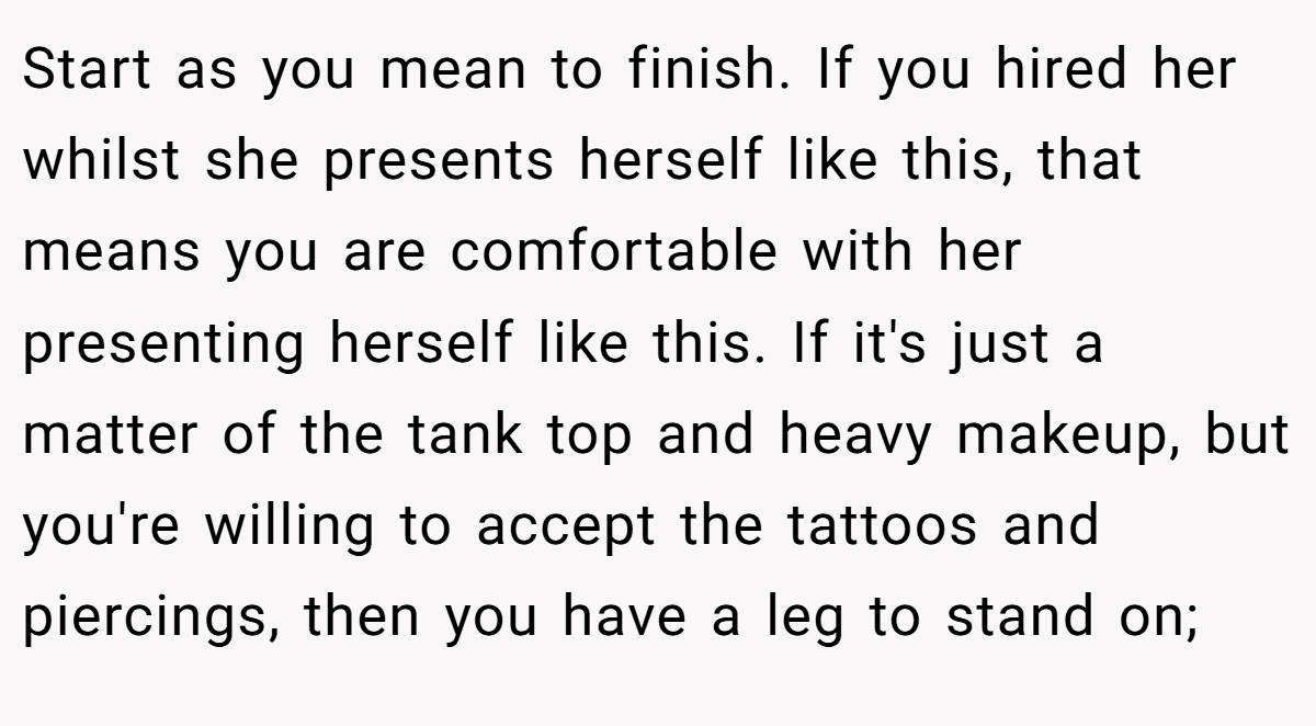 Start as you mean to finish. If you hired her whilst she presents herself like this, that means you are comfortable with her presenting herself like this. If it's just a matter of the tank top and heavy makeup, but you're willing to accept the tattoos and piercings, then you have a leg to stand on;